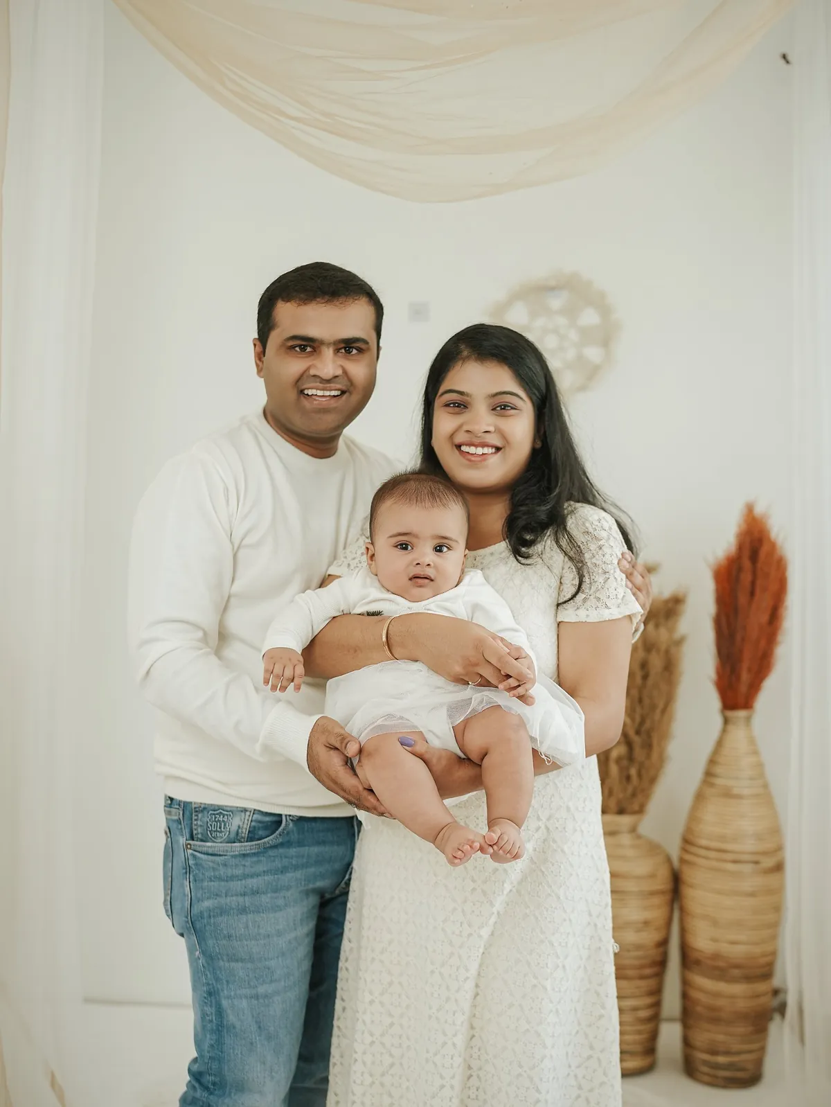 Smiling parents in white hold their baby between them in a bright room with draped fabric and pampas grass.