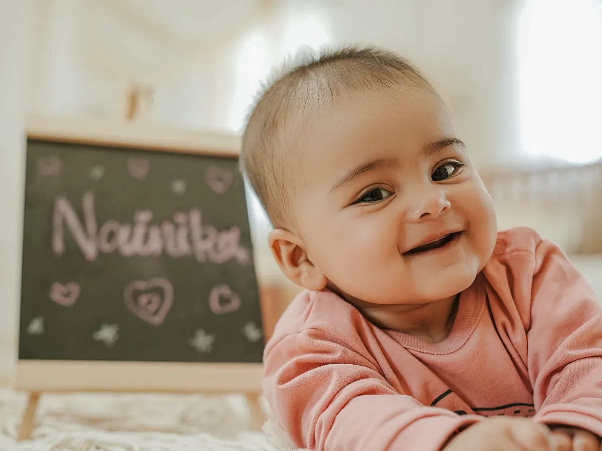 A baby in a pink top grins at the camera during tummy time, with a small chalkboard in soft focus behind.