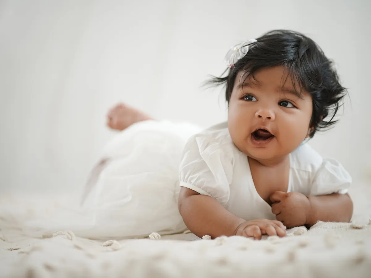 A baby with thick dark hair and a white dress lifts her head during tummy time on a cream blanket, mouth open in delight.