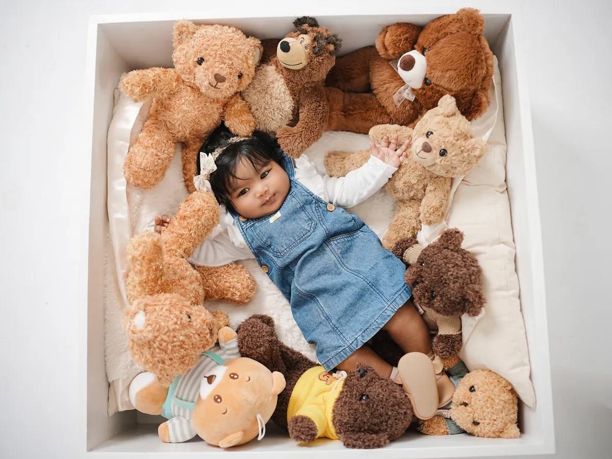 Seen from above, a baby in a denim pinafore lies in a white box surrounded by teddy bears.