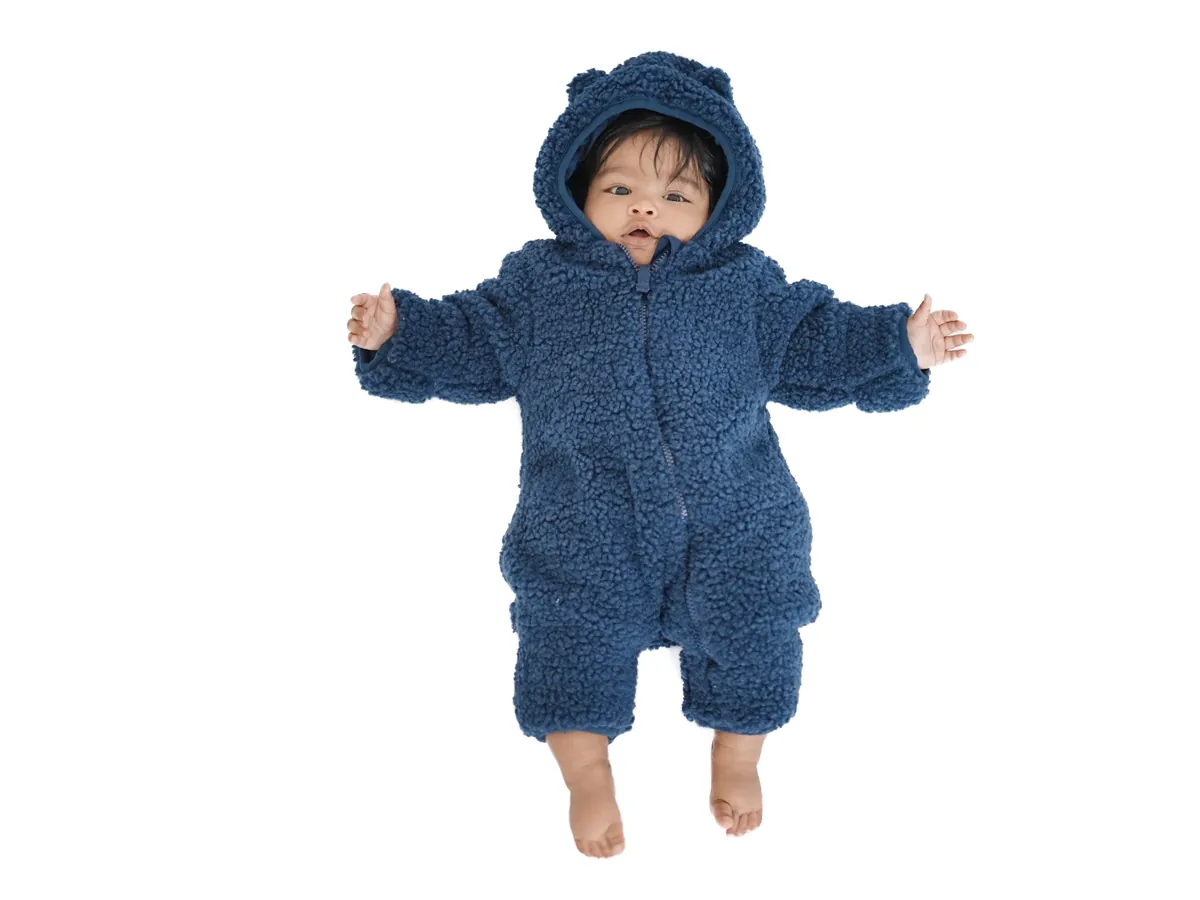 Seen from above, a baby in a navy fleece bear suit lies with arms outstretched on a pure white background.