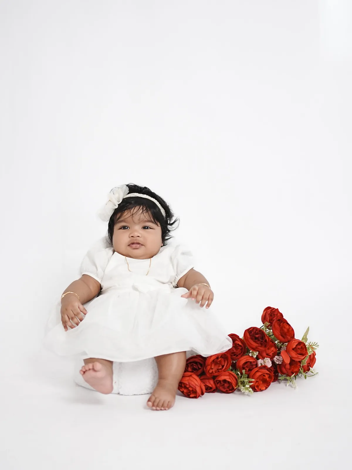 A baby in a white dress and headband sits propped on a white backdrop beside a spray of red roses.