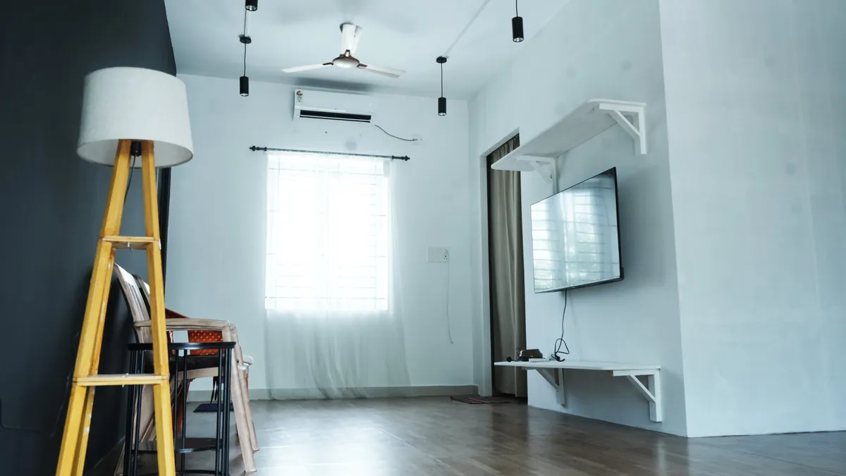 A small lounge with a floor lamp, chairs, a wall-mounted screen, an air conditioner and a curtained window.